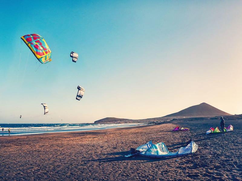 Plage avec kitesurfeurs colorés et mer calme sous un ciel clair, collines en arrière-plan.