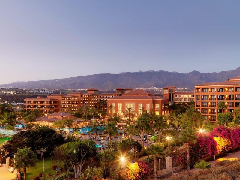 Groot hotelresort met bergen op de achtergrond bij schemering, omringd door groen.