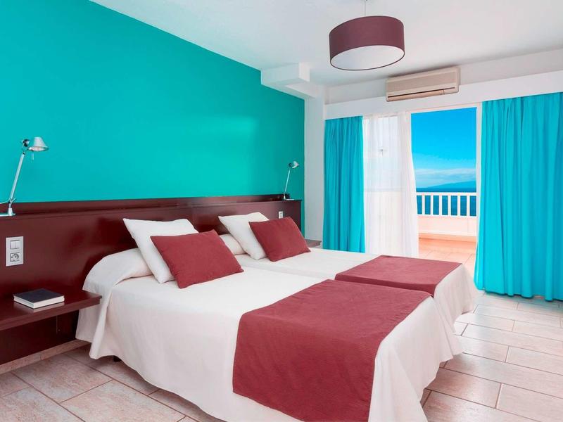 Habitación de hotel luminosa con cama doble, pared turquesa y balcón con vista al mar.