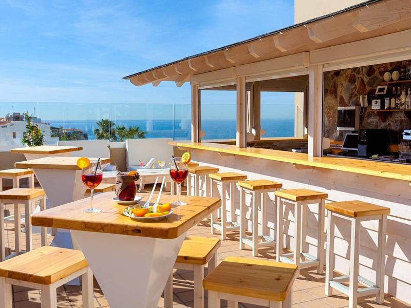 Bar al aire libre con taburetes y bebidas en una terraza soleada con vista al mar.