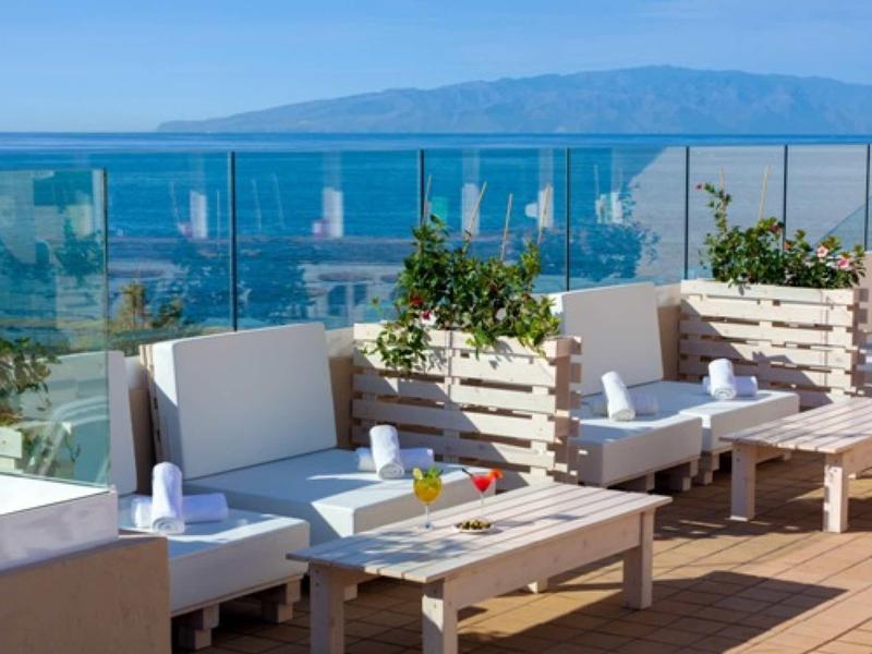 Terraza con muebles blancos de salón, suelo de madera y vista al mar al fondo.
