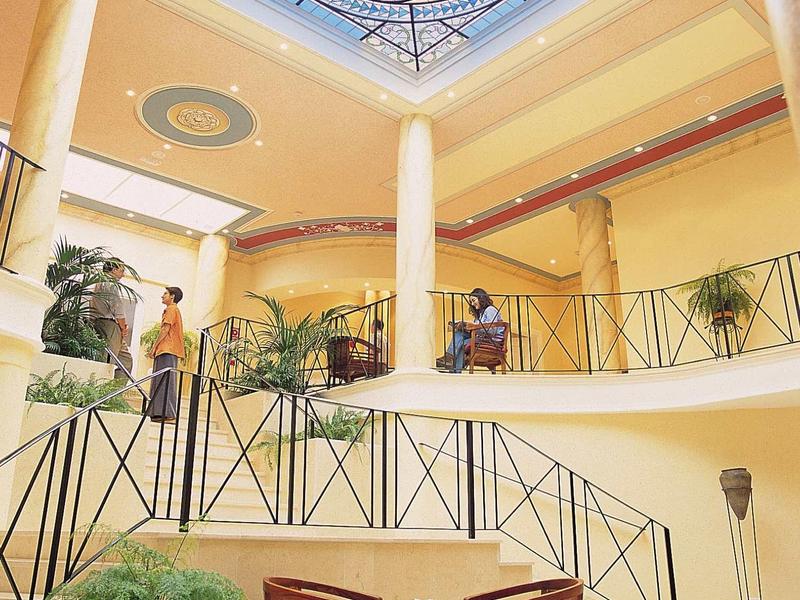 Hell erleuchteter Innenraum mit Glasdach, Pflanzen, Säulen und Tisch mit zwei Stühlen.