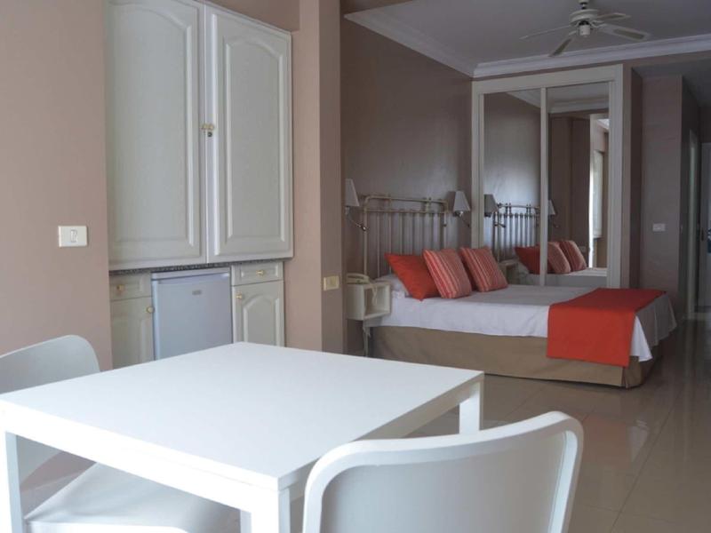 Helles Zimmer mit weißem Esstisch, zwei Stühlen, Bett mit orangefarbenen Kissen und Spiegelkleiderschrank.