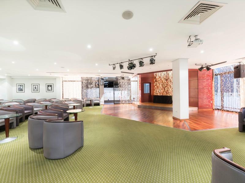 Ampia sala eventi luminosa con moquette verde, gruppi di sedute e palco.