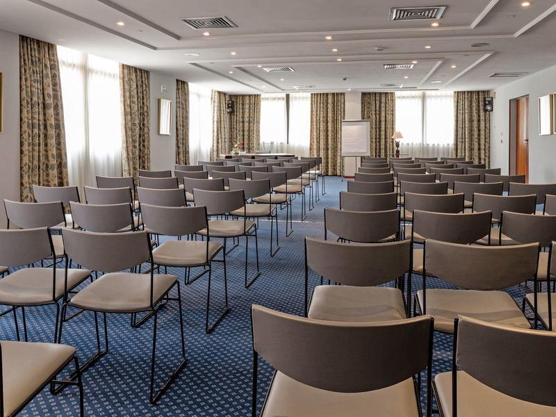 Sala conferenze con file di sedie e grandi finestre, atmosfera luminosa e moderna.