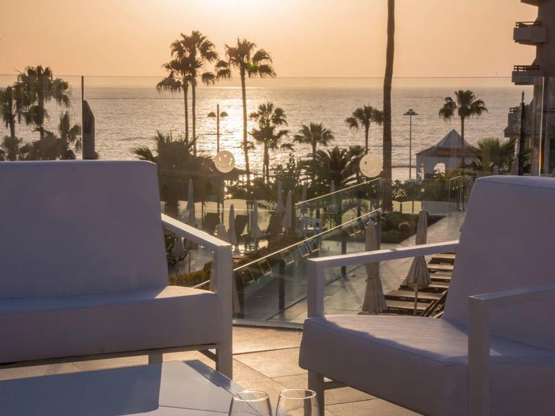 Zwei Sessel und ein Tisch mit zwei Gläsern vor Sonnenuntergang am Pool mit Palmen und Meer.