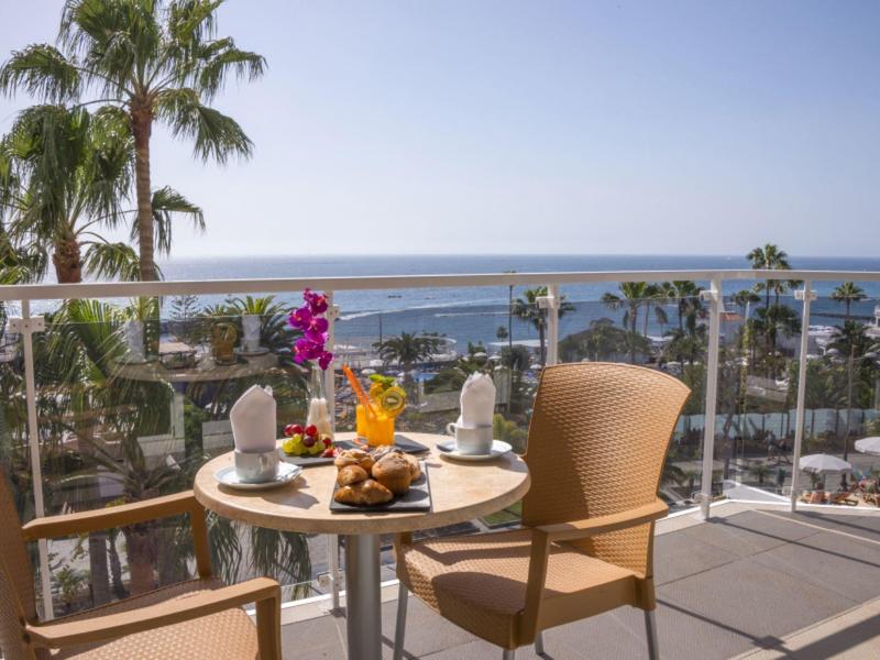 Balkontisch mit Frühstück, Palmen und Meer im Hintergrund bei klarem Himmel an sonnigem Tag.