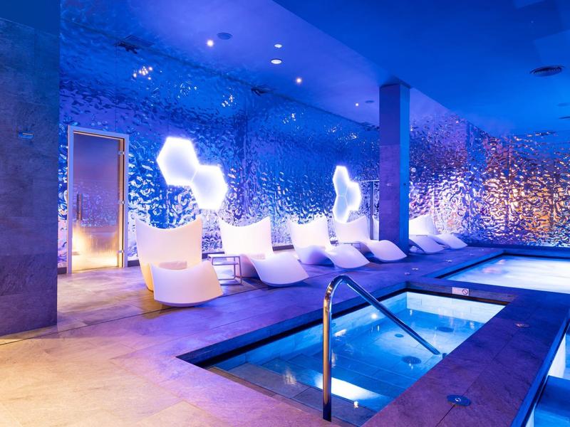Modernes Innenhallenbad mit Whirlpool und weißen Liegen vor blau beleuchteter Wand