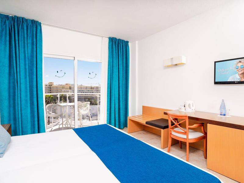 Helles Hotelzimmer mit Bett, blauem Läufer, Schreibtisch und Blick auf Stadt durch Fenster mit blauen Vorhängen.