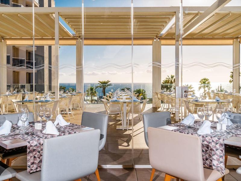 Elegantes Restaurant mit Blick auf das Meer, helle Möbel und vorbereitete Tische unter Holzpergola.