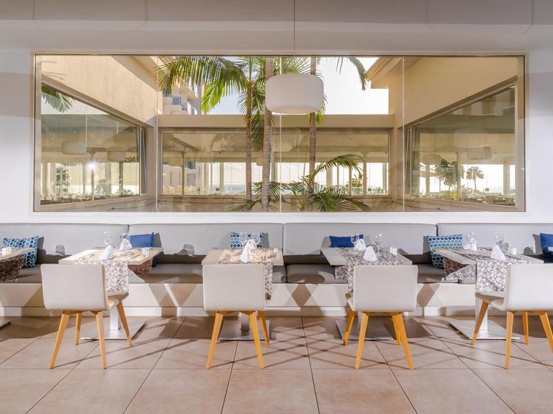 Moderner Außenbereich mit weißen Sesseln und Holztischen vor großen Fenstern und Palmen.