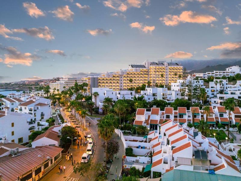 Städtische Urlaubsszene mit weißen Gebäuden, belebter Straße und Bergkulisse bei Sonnenuntergang.