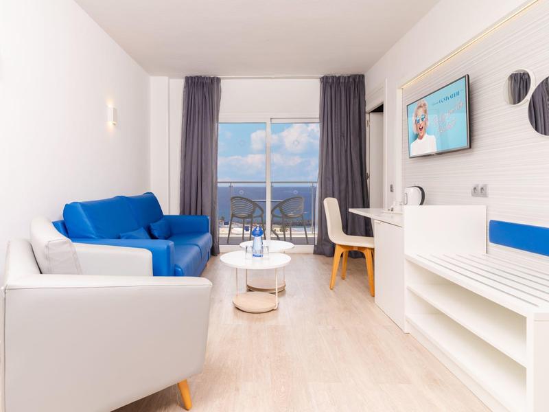 Modern eingerichtetes Hotelzimmer mit blauem Sofa, Schreibtisch und Meerblick durch großes Fenster