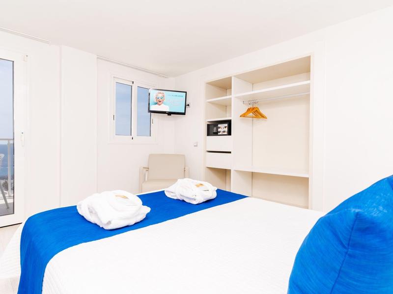Helles Hotelzimmer mit Doppelbett, blauer Bettwäsche, offenem Schrank und kleinem Fernseher.