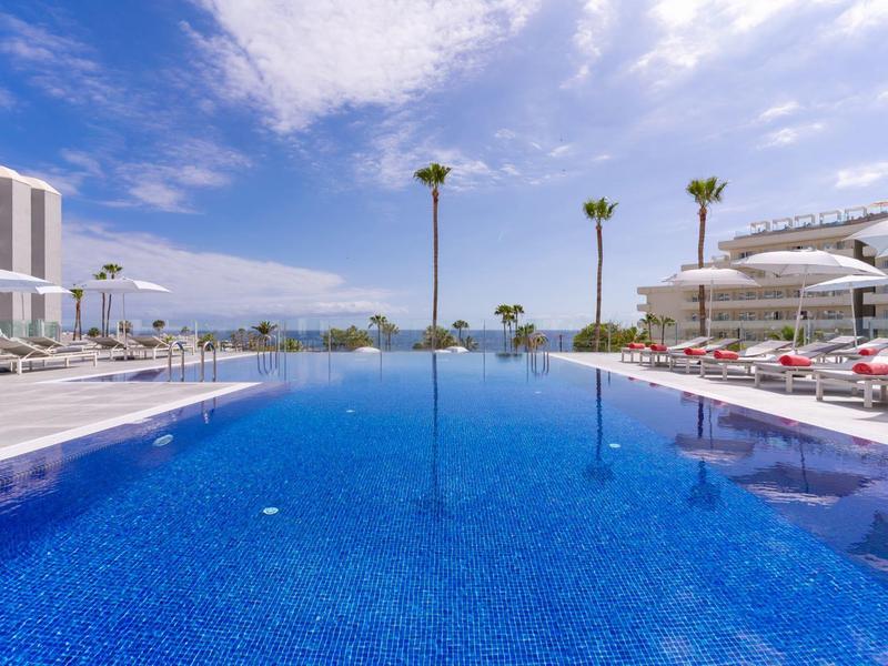 Großer Außenpool mit klar blauem Wasser, umgeben von Liegestühlen und modernen Gebäuden bei sonnigem Himmel.