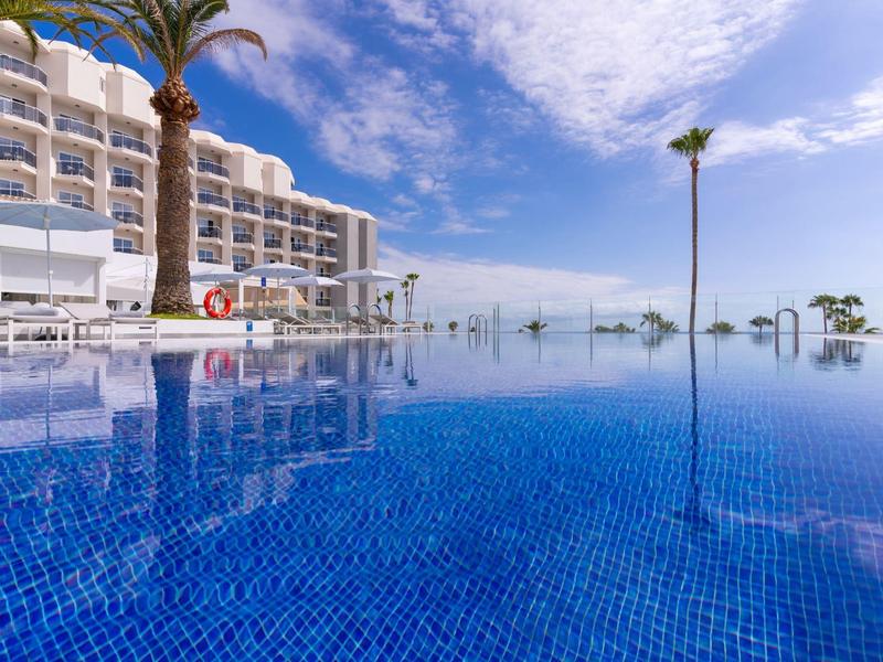 Schwimmbecken mit Palmen und Hotelgebäude unter blauem Himmel mit Wolken.