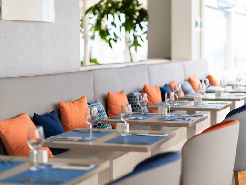 Moderner Essbereich mit Sitzbänken, orangenen Kissen und eingedeckten Tischen in einem hellen Raum.