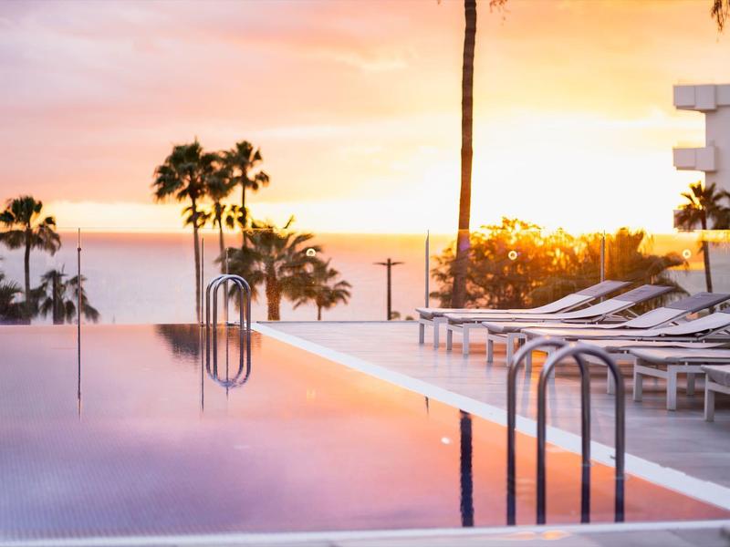 Infinity-Pool mit Liegestühlen und Palmen bei Sonnenuntergang am Meer