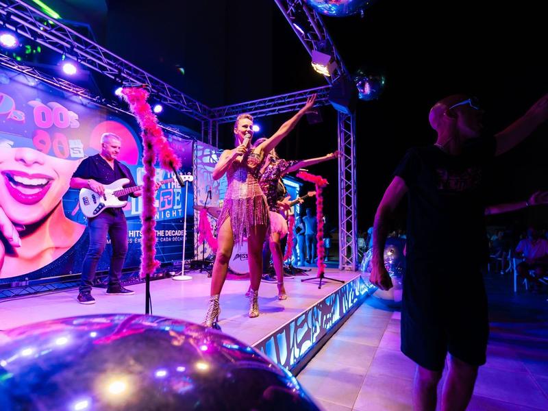 Bühnenauftritt mit Sängern, Gitarrist und Tänzern in lebhafter Clubatmosphäre.