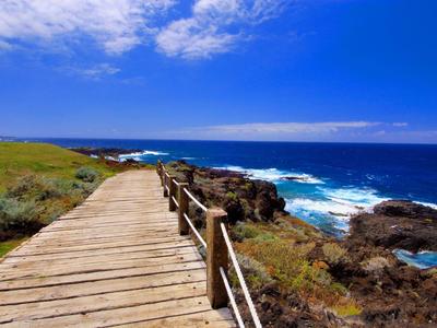 Drewniany pomost biegnie wzdłuż wybrzeża z widokiem na ocean i częściowo pochmurne niebo.