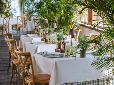 Jasna restauracja z stołami przykrytymi białymi obrusami i wieloma zielonymi roślinami.