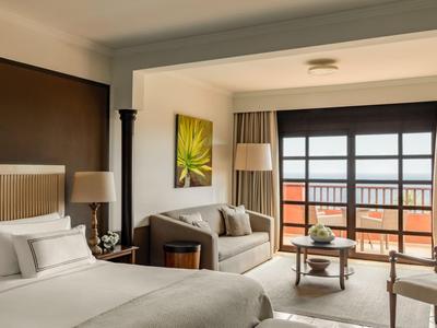 Brightly furnished hotel room with double bed, sofa, and balcony with sea view.