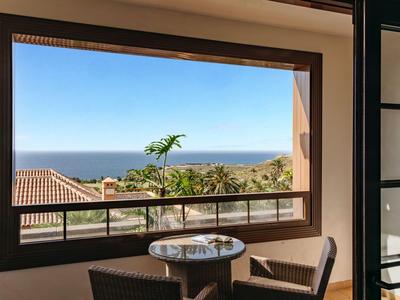 View through a window of the sea, with a table and two chairs on the balcony.