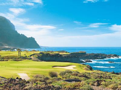 Zielony pole golfowe z piaskownicą przy morzu pod niebieskim niebem z górami w tle.