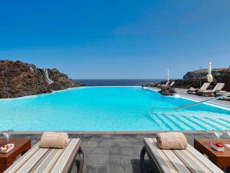 Basen infinity z leżakami na czarnej kamiennej platformie z widokiem na morze pod czystym niebem.