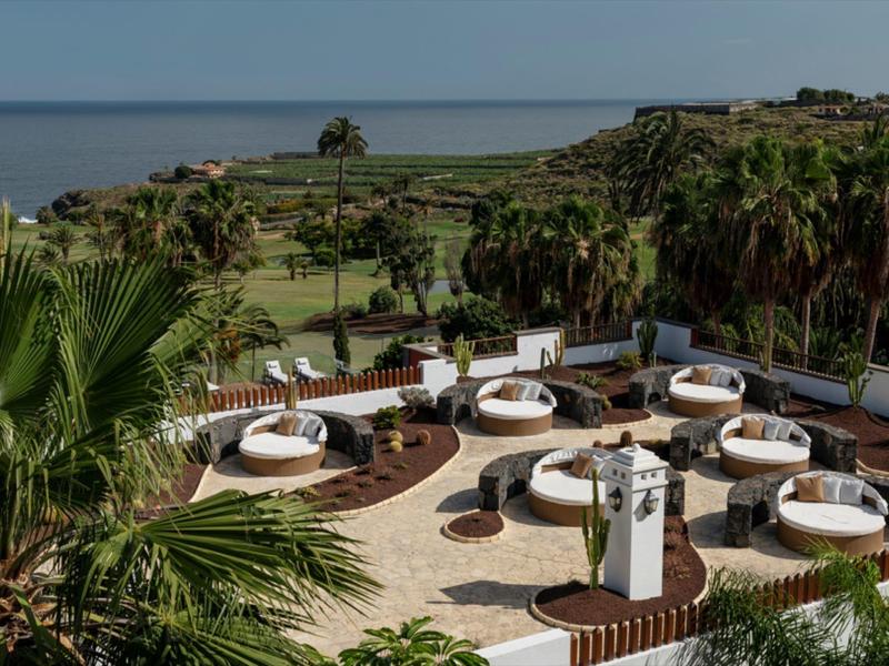 Taras z okrągłymi miejscami do siedzenia otoczony palmami z widokiem na morze.