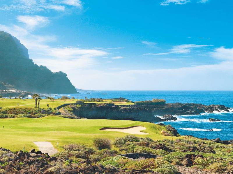 Zielony pole golfowe z piaskownicą przy morzu pod niebieskim niebem z górami w tle.