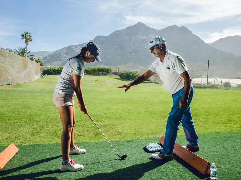 Dwie osoby grające w golfa na polu treningowym z górami i czystym niebem w tle.