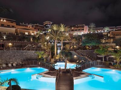 Nächtlicher Blick auf einen beleuchteten Pool mit Brücke und Palmen in einem Resort.