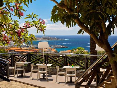 Terrasse ombragée avec tables et chaises face à la mer et aux arbres fleuris.
