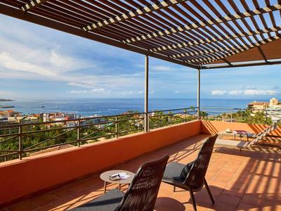 Terrasse ensoleillée avec vue sur la mer, fauteuils noirs et table sous pergola ombragée.