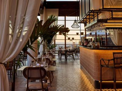 Coin bar élégant avec tabourets noirs, grandes plantes et rideaux blancs légers.