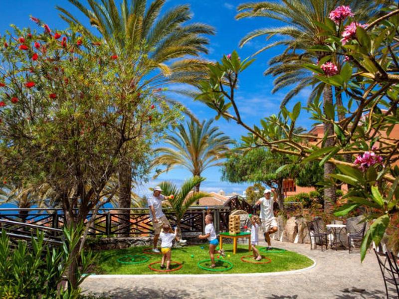 Jardin luxuriant avec palmiers, enfants jouant et tables en plein air sous un ciel bleu vif.