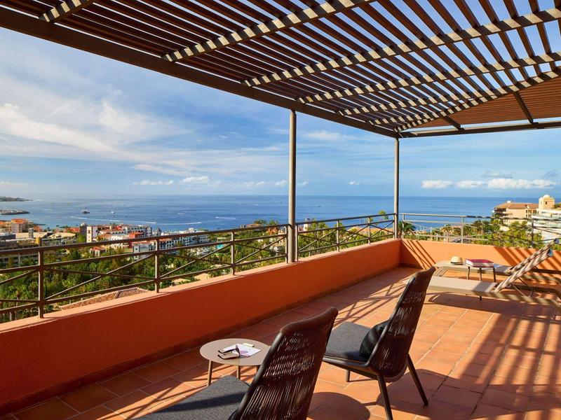 Terrasse ensoleillée avec vue sur la mer, fauteuils noirs et table sous pergola ombragée.