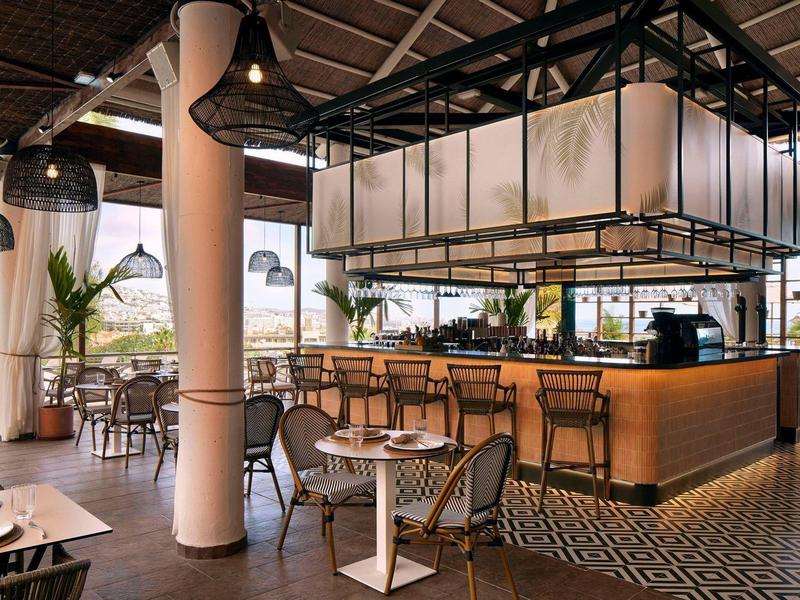 Moderní restaurace s barem, dřevěnými stoly a židlemi ve světlém prostoru.