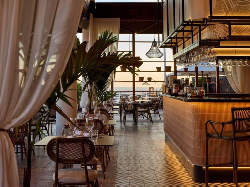 Coin bar élégant avec tabourets noirs, grandes plantes et rideaux blancs légers.