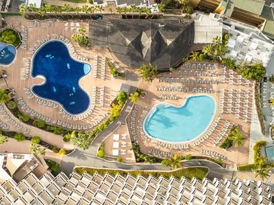 Aerial view of two swimming pools with sun loungers surrounded by palm trees at a hotel resort.