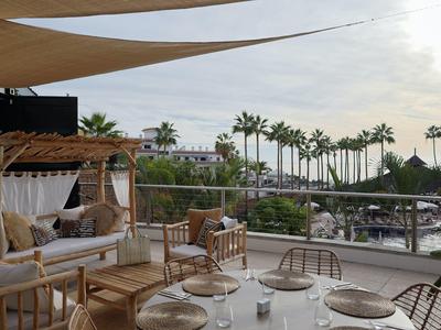 Cozy terrace with wooden furniture, cushions, and sea view under cloudy sky.