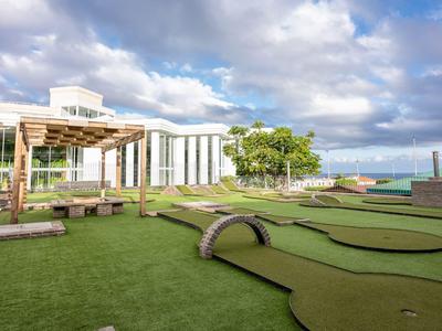 Pole do minigolfa z trawą i nowoczesnym hotelem w tle pod pochmurnym niebem.