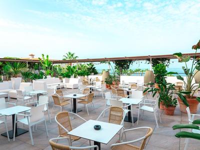 Taras na dachu z białymi stołami, krzesłami i wieloma zielonymi roślinami pod niebieskim niebem.