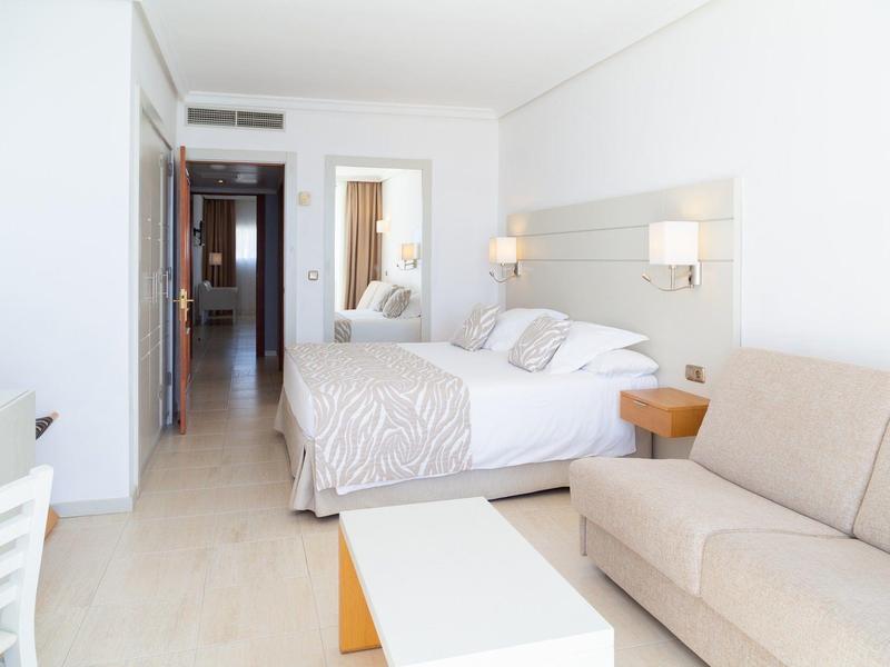 Bright hotel room with double bed, beige sofa, white table, and corridor leading to bathroom.