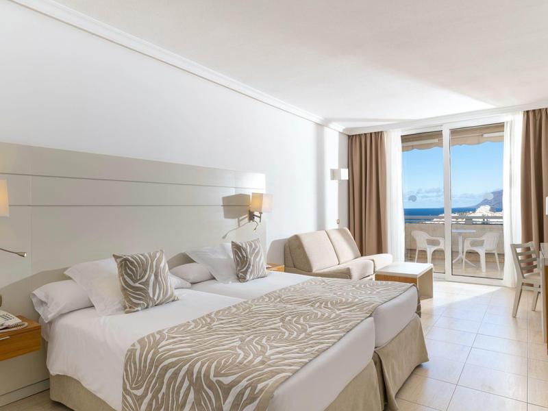 Bright hotel room with two beds, seating area, and balcony with sea view.