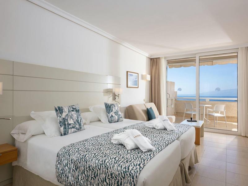 Bright hotel room with double bed, seating area, and sea view through large windows.