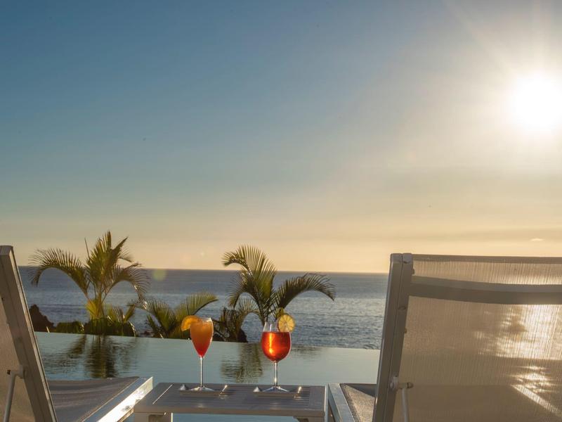 Two lounge chairs with drinks on a table by an infinity pool at sunset.