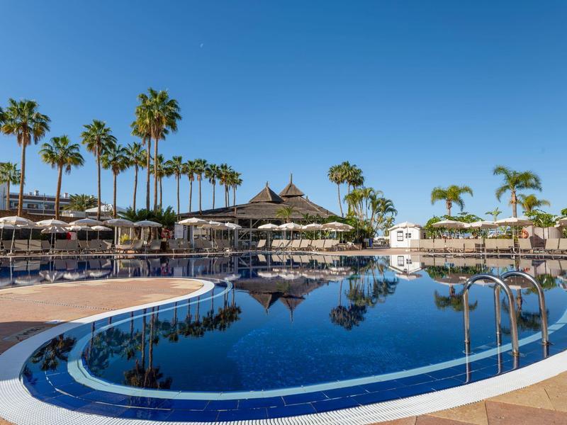 Clear blue pool area with palm trees, loungers, and umbrellas under a blue sky.