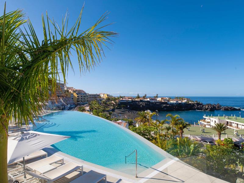 Infinity pool with lounge chairs, palm trees, and a view of the sea and harbor under a clear blue sky.